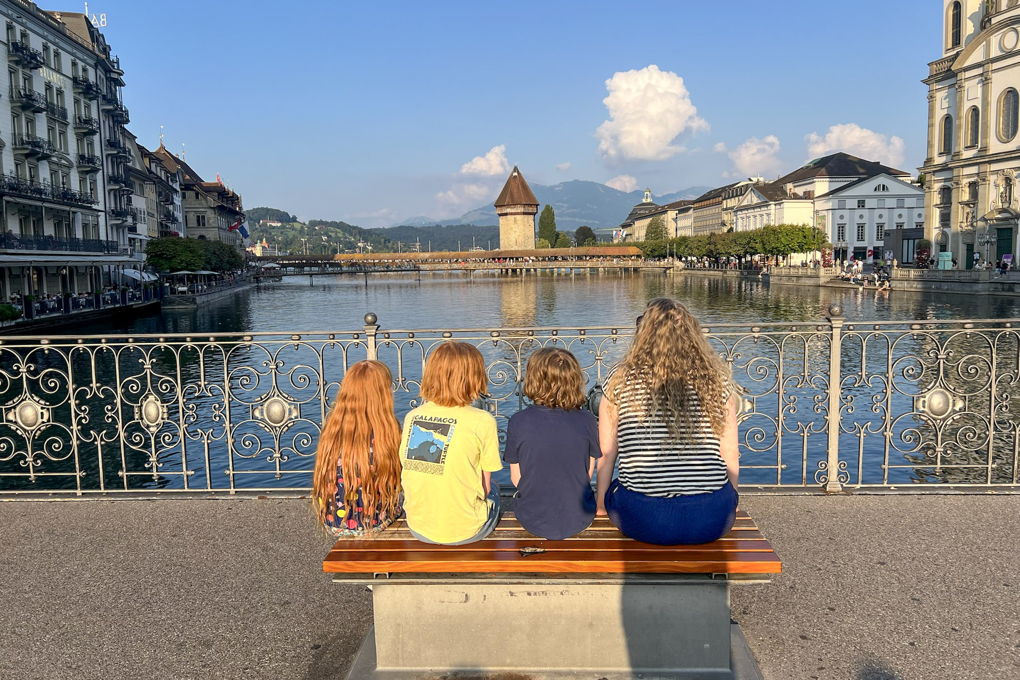 Three children and an adult sit on a bench overlooking the river at Lucerne.